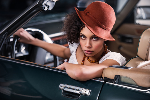 Fashionable retro African American woman is sitting in an old timer car and looking at the camera.