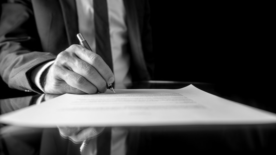 Black and white low angle image of the hand of a businessman in a suit signing a document or contract with a fountain pen on a reflective surface.