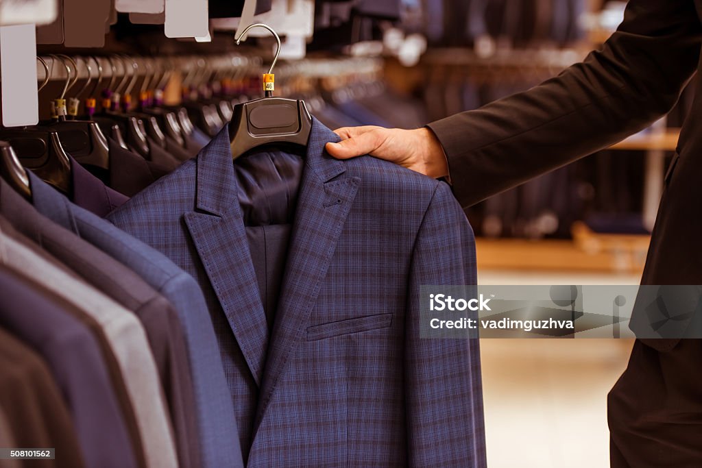 People in suit shop A hand of a modern young handsome businessman choosing classical suit from the row of suits in the suit shop, close-up Menswear Stock Photo