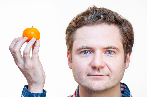 Portrait of young, blue-eyed, elegant, Caucasian Ethnicity, holding tangerine in right hand near his head on white background. Creative thinking and nonstandard solutions.