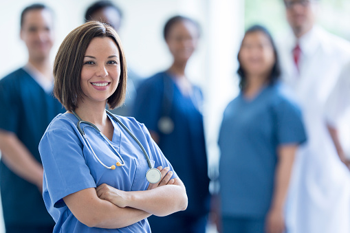 A nurse is happily standing in front of her colleagues at the hospital. She is wearing her scrubs and is smiling while looking at the camera.