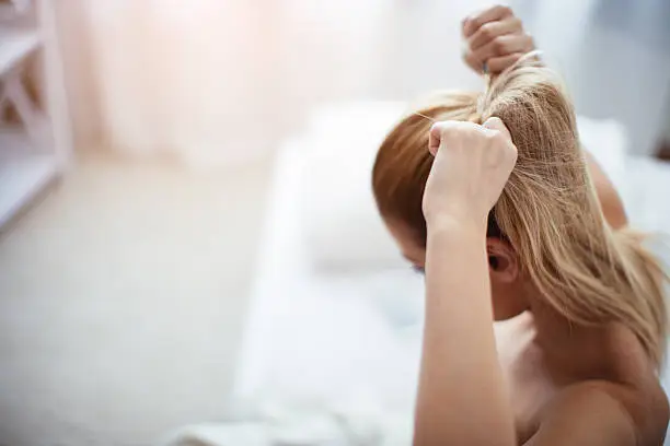 Young woman sitting at the bed in her bedroom, getting ready for work, tying her hair.