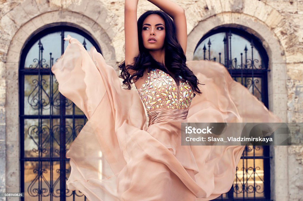 beautiful woman with dark hair in luxurious silk dress fashion photo of beautiful glamour model with long dark hair  in luxurious silk beige dress posing on castle background Adult Stock Photo