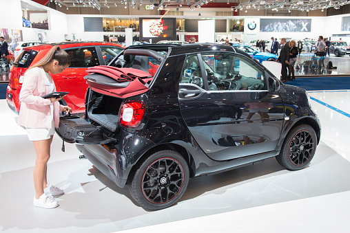 Brussels, Belgium - Januari 12, 2016: Smart ForTwo Cabrio compact convertible city car rear view. The Smart Fortwo is a rear-engine, rear-wheel-drive, 2-seater small hatchback city car produced by smart Automobile, a division of Daimler AG. A girl is checking the boot of the car. The car is on display during the 2016 Brussels Motor Show. The car is displayed on a motor show stand, with lights reflecting off of the body. There are people looking around and other cars on display in the background.