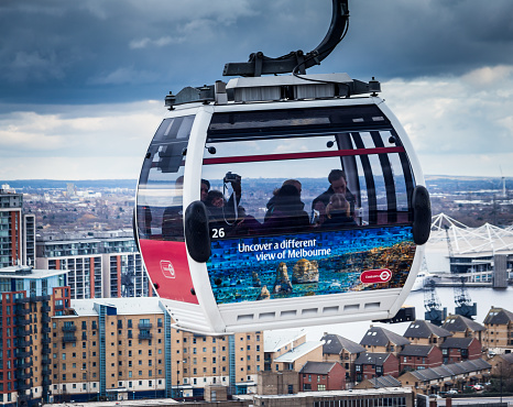London, UK - February 21, 2015: An Emirates Airline cable car ascends after departing the Royal Docks terminus in Greenwich, London, on the north side of the River Thames. The cable car is filled with tourists who are staring out at the urban panorama - tower blocks and other residential structures - below. The winter's day is overcast and an ominous sky is brooding overhead.