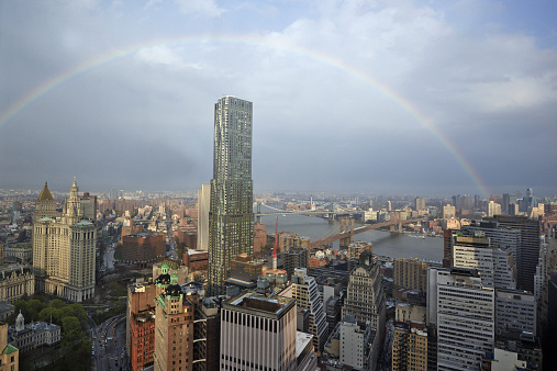 A rainbow forms over Lower Manhattan, the Brooklyn Bridge, the Manhattan Bridge, East River and professional office buildings on a spring day.