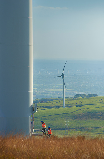 two wind farm engineers walk toward the door way of a wind turbine. they are wearing orange hi vis jackets and blue hard hats . one is male , one is female. In the background wind turbines can be seen across the landscape down to a distant city .