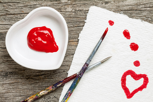 Heart-shaped porcelain bowl with glob of red paint on a wooden surface with artist´s brushes painting a heart on hand-made paper. Heart to heart.