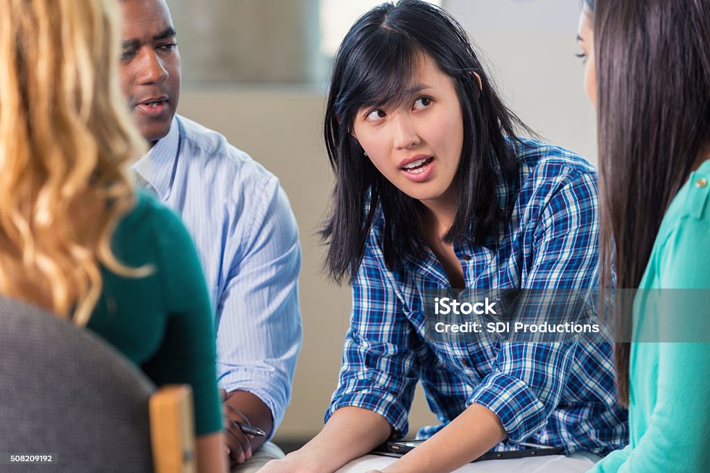 Young Asian woman expresses concern in support group Serious young Asian woman talks about her problems in group therapy or support group. She has a concerned expression on her face as the talks to the group. A diverse group of patients listen to her talk. She is wearing a blue plaid shirt. Psychotherapy Stock Photo