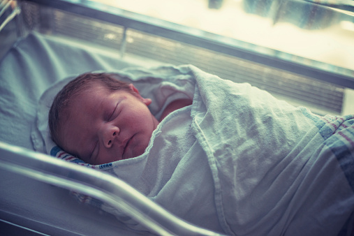 A baby boy sleeps in a maternity ward bassinette, wrapped tightly in a blanket.
