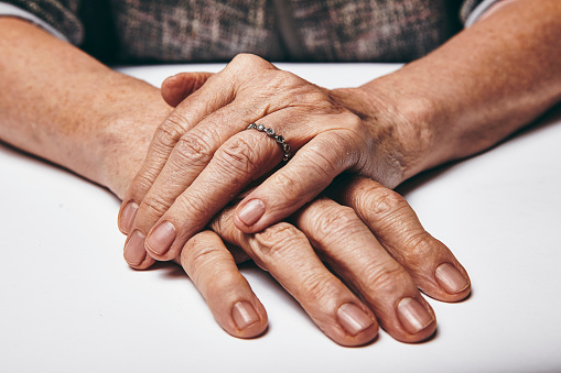 Macro of an old lady sitting with her hands clasped on a table. Elderly woman's hands with a ring resting on grey surface. Focus on hands.