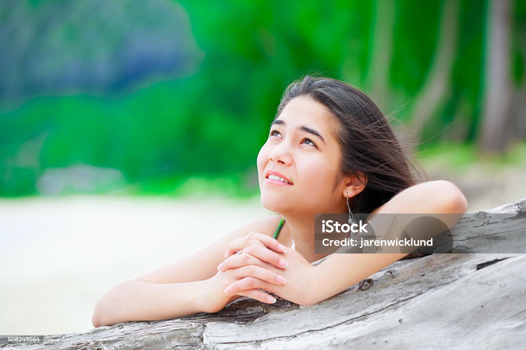 Beautiful teen girl on beach praying by driftwood log Beautiful biracial  teen girl on tropical  beach praying by driftwood log Contemplation Stock Photo