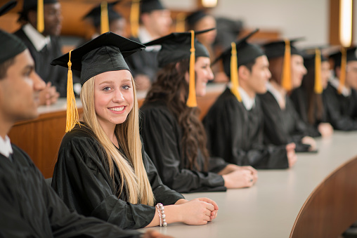 A multi-ethnic group of college graduates are sitting together in a lecture hall and are listening to their professor. They are wearing a cap and gown and one woman is smiling and looking at the camera.