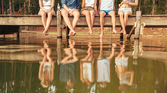 Legs of young people sitting on the edge of a jetty hanging down to the water. Group of friends hanging out at the lake.