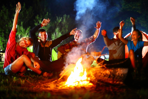 Group of  mid 20's friends sitting by campfire at remote location outside the city.They are singing and laughing.All waving and looking at camera.There are two girls and three guys.