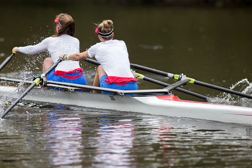 Two teenage girls are training for their rowing team on the river in a double scull boat. Rear view of them rowing through the water down river.