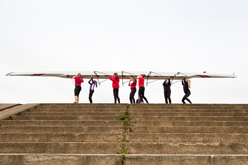 Teenage girls standing at the top of some steps carrying a skull boat ready to train on the river. View from the bottom of the stone steps looking up.