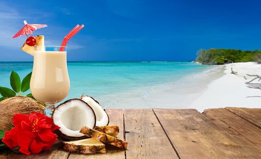 Glass of refreshing piña colada on wood table shot against tropical beach background. The glass has two red drinking straws, a pineapple slice garnished with maraschino cherry and a pink umbrella. At the base of the glass are two halves of coconut, a whole coconut, three slices of pineapple and a red hibiscus flower. The foreground composition is at the left of the frame leaving a useful copy space at the center-right.