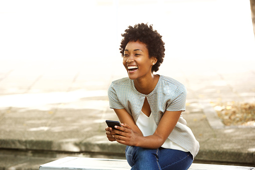 Portrait of beautiful young african woman smiling while sitting outside on a bench holding mobile phone