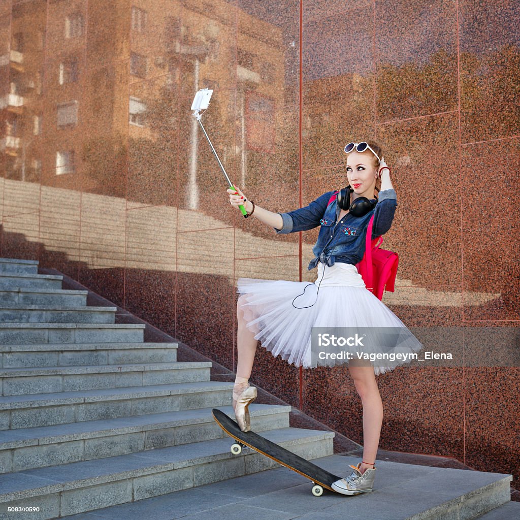 Ballerina hipster. Selfie on the street. Girl listening to music on headphones. Cute ballerina makes hipster selfie phone on selfie stick. A girl wearing tutu, ballet shoes and sneakers, sunglasses. Girl riding on skateboard. The concept of youth fashion. Arts Culture and Entertainment Stock Photo