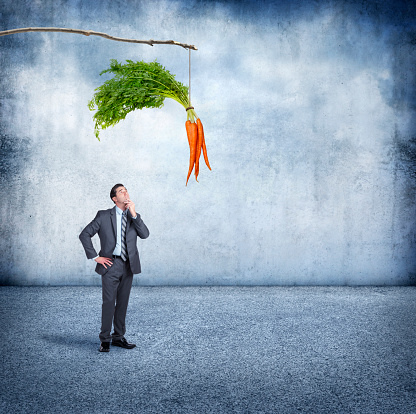 A businessman places his hand on his chin as he looks up at a bunch of carrots that are dangling from a stick directly above him.  He is pondering what it will take to motivate him.