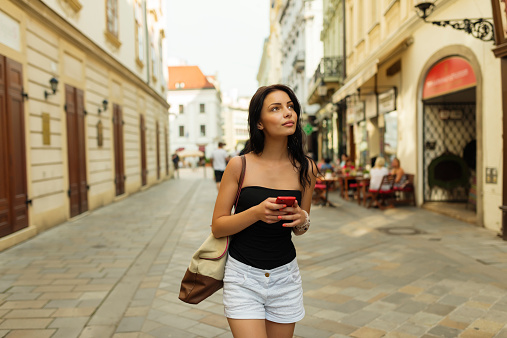 Teen woman with black hair and in black clothing with travel bag is traveling, looking and taking pictures with her red smart phone in historical part of Bratislava city  in hot summer afternoon. Shallow DOF. There is street  with shop windows and patio in the background. Whole scene is illuminated by soft daylight. Low depth of field and blurred background.