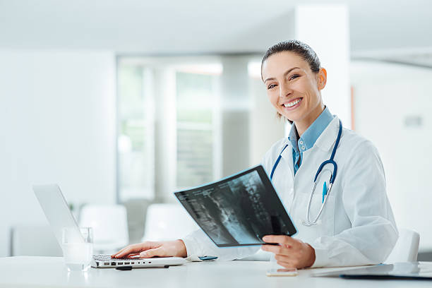 Smiling female doctor examining an x-ray Smiling confident female doctor sitting at office desk and examining a patient's x-ray, she is looking at camera examining x ray stock pictures, royalty-free photos & images