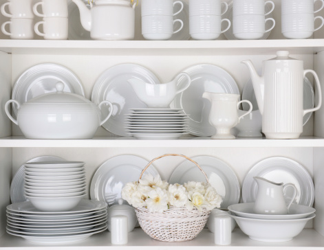 Closeup of white plates and dinnerware in a cupboard. A basket of white roses is centered on the bottom shelf. Items include, plates, coffee cups, saucers, soup tureen, tea pot, and gray boats.