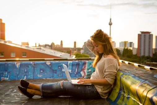 young woman sits on roof, works on laptop - back lit