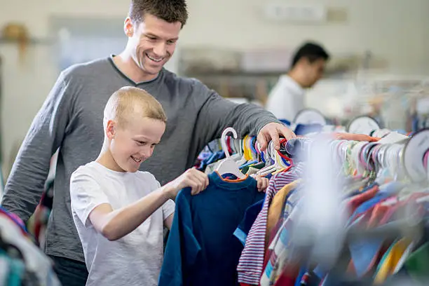 A little boy and his father are looking through a rack of clothes while shopping together. The boy has pulled out a long sleeve shirt to buy.