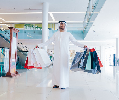 hero young Arab man with traditional Emirati clothes carrying a lot of shopping bags and smiling at modern shopping mall