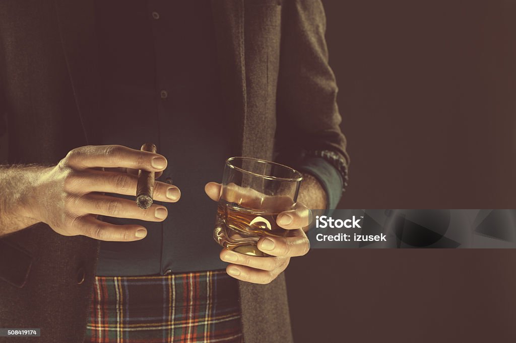 Man drinking whiskey and smoking cigar, close up of hands Elegant man wearing tweed jacket and tartan skirt, holding glass of scotch whisky and cigar. Close up of hands, unrecognizable person. Dark tone, black background. Whiskey Stock Photo