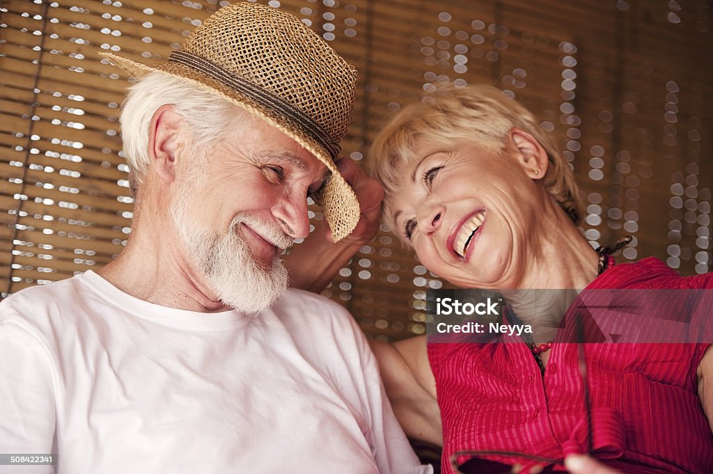 Cheerful Senior Couple Enjoying Summer Day on a Terrace Senior couple enjoying the moment on the summer terrace. She has blond short hair and he has gray hair and beard. He is wearing the straw hat and she red silk blouse. They are smiling to each other. 60-69 Years Stock Photo