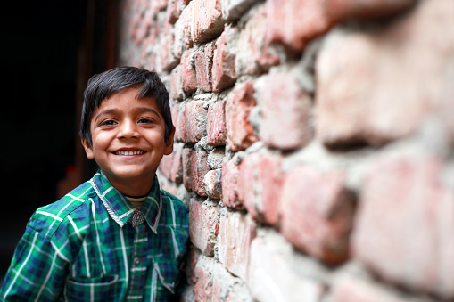 Cheerful little poor boy standing portrait near brick wall at his home and looking to the camera portrait close up. 