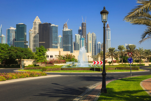 A typical example of urban landscaping in the modern Arabian city of Dubai, in the United Arab Emirates.  In the background are skyscrapers of the city and some construction is still going on.  In the foreground, date palms, flowering shrub and lawns. In the centre of the photo is a roundabout with a fountain. The morning sun throws long shadows across the road.  It is hard to believe that this is the Arabian desert; every blade of grass is artificially grown. Photo shot in the morning sunlight; horizontal format. Copy space. No people.