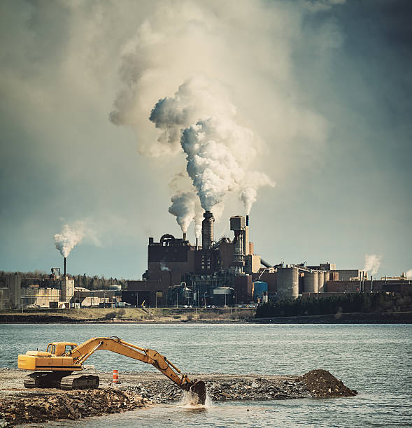 An excavator works to infill a harbour while a factory grinds away in the distance.