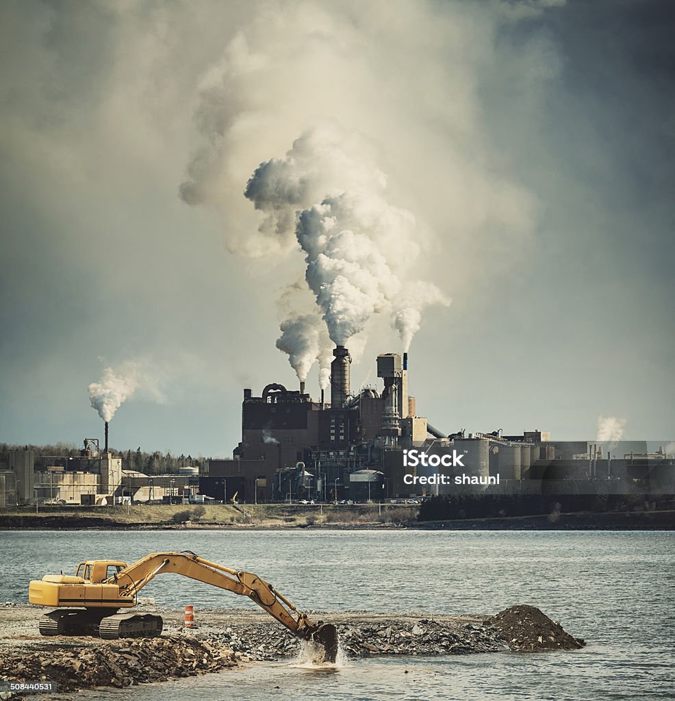 The Drone of Industry An excavator works to infill a harbour while a factory grinds away in the distance. Pollution Stock Photo