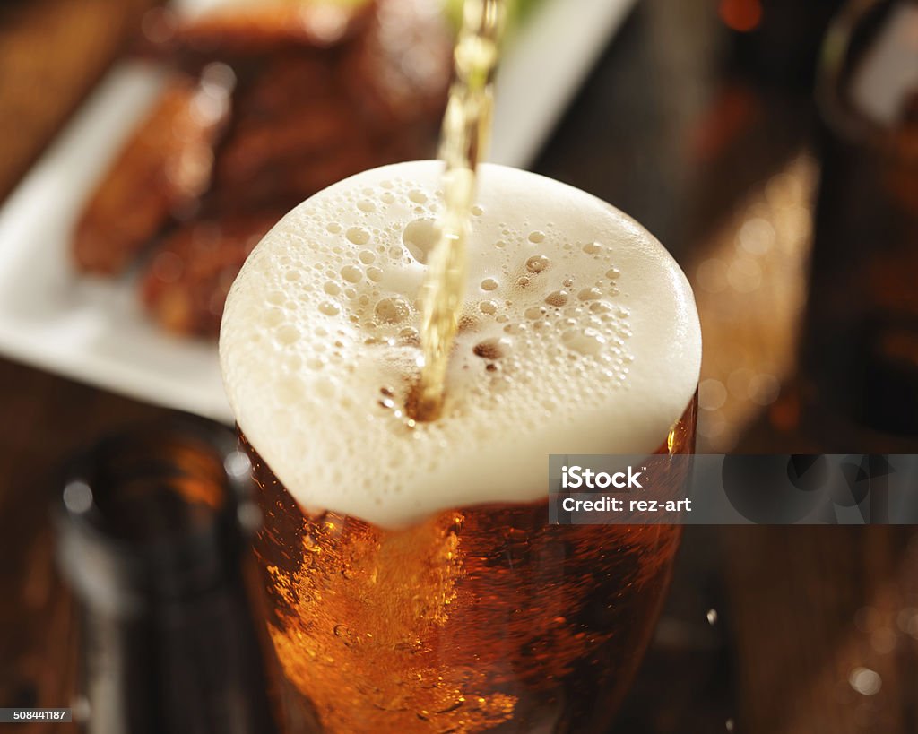 pouring beer into glass with foam pouring beer into glass with bbq chicken wings in background Beer - Alcohol Stock Photo