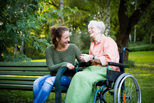 charming young woman and senior woman in a  wheelchair sit together in a park and have fun