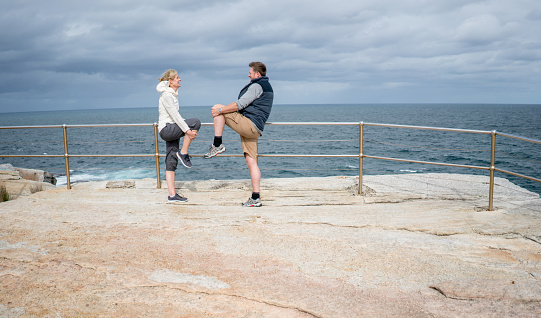 Couple exercising together outdoors by the beach stretching their legs and looking very happy