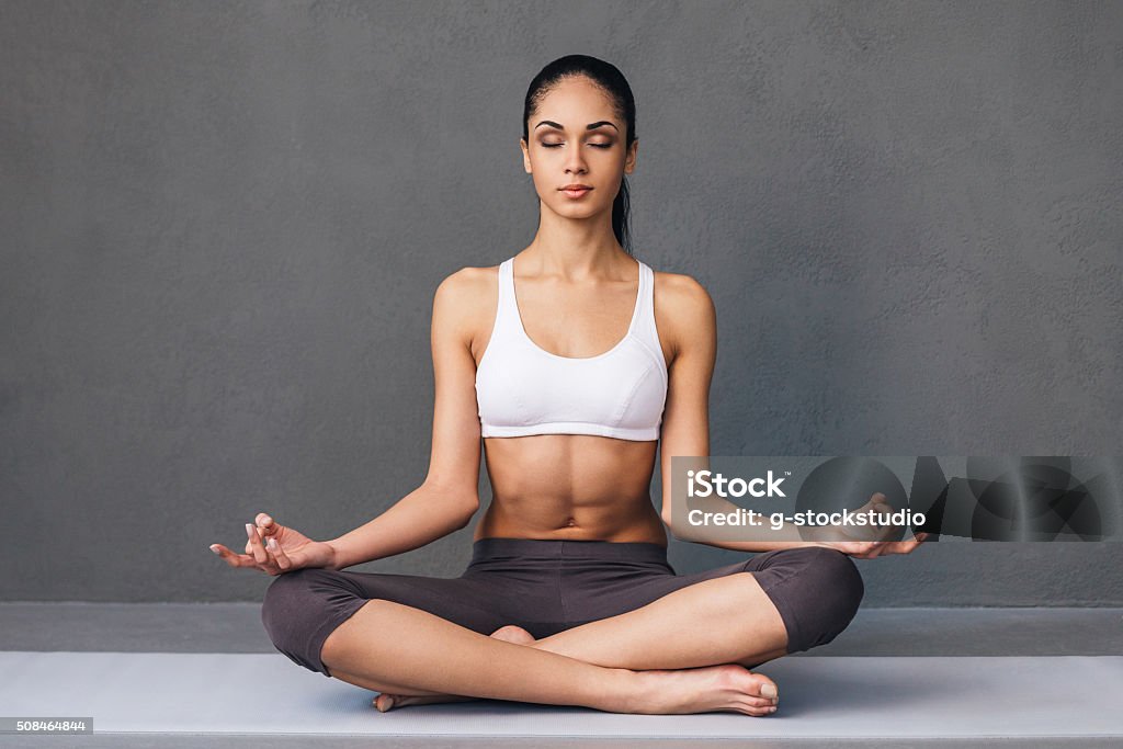 Long breath in. Beautiful young African woman in sportswear practicing yoga while sitting in lotus position against grey background Yoga Stock Photo