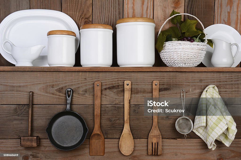 Rustic Kitchen Closeup of a rustic kitchen wall. One shelf with canisters, plates and a basket. Hanging on the wall below are wooden utensils, frying pan and towel. Kitchen Stock Photo