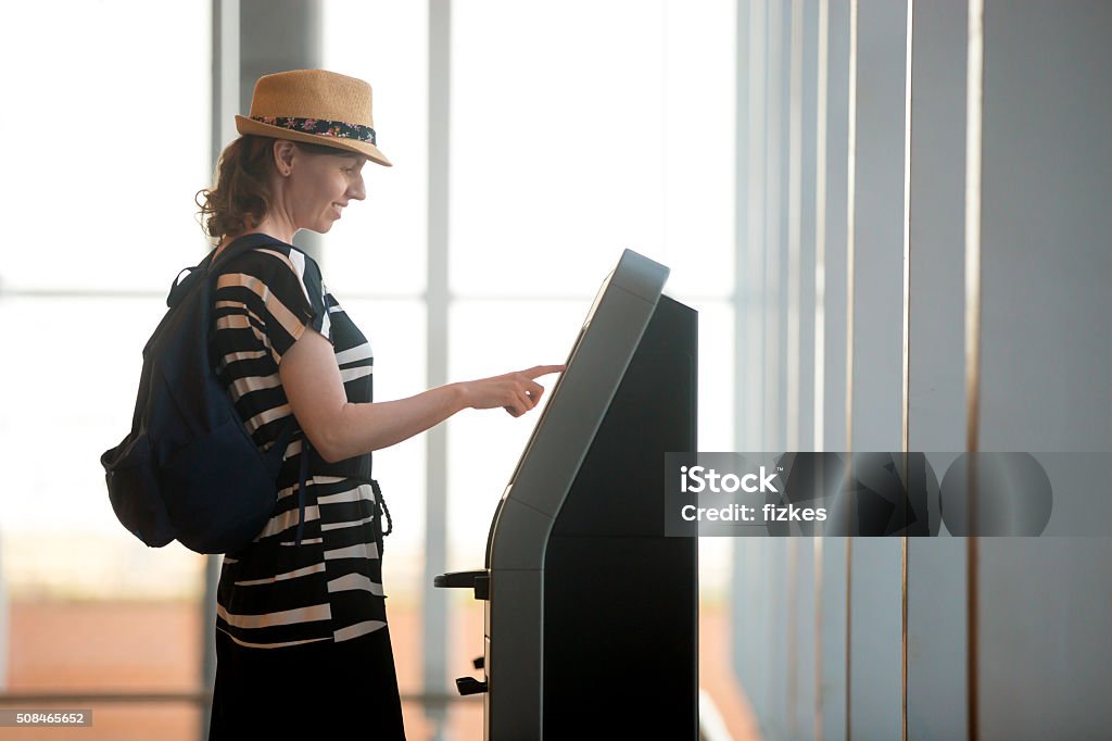 Woman doing self-check-in in airport Young woman at self service transfer area doing self-check-in or buying plane tickets at automated machine with touchscreen interactive display in modern airport terminal building Kiosk Stock Photo