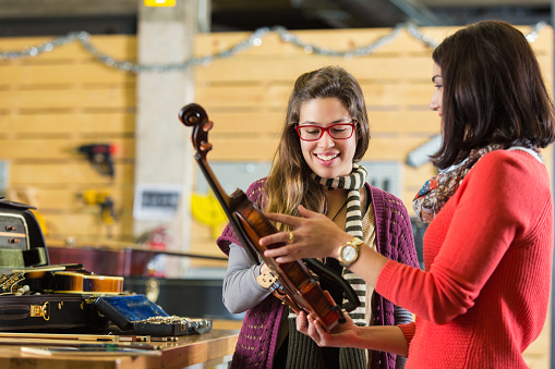Professional violinist is brining her violin into musical instrument repair shop to be repaired. She hands her violin to young female employee. The women talk about the repair. Musical insturments and tools are on the table beside them.