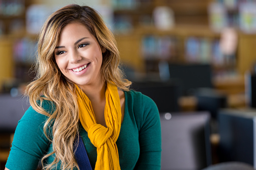 Pretty Hispanic high school or college student is standing in school library. Desktop computers and bookshelves are behind her. She has long light brown hair and is wearing a yellow scarf.