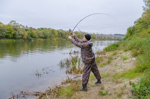 The fisherman in a camouflage catches fish on the river a spinning.