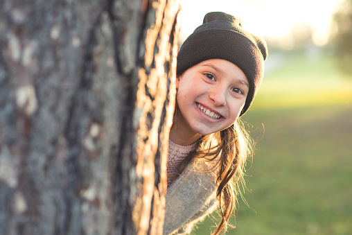 Portrait of a little girl hiding behind a tree in a park. She is smiling and looking at camera. Sun is shining from behind.