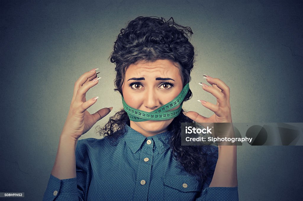 Diet restriction stress. Frustrated woman with measuring tape around mouth Diet restriction and stress concept. Portrait of young frustrated woman with a green measuring tape around her mouth isolated on gray wall background. Face expression emotion Addiction Stock Photo