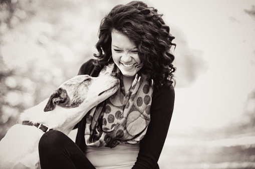 A teen girl laughing and smiling as her dog snuggles up to her to nuzzle her with his nose. Girl is African American. Both are outdoors. High resolution color photograph processed in vintage black and white with a hint of sepia tones. Horizontal composition.