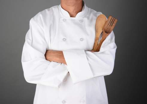 Closeup of a chef with his arms folded holding wood utensils. Man is unrecognizable. Horizontal format on a light to dark gray background. Model Released.